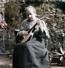 Self Portrait Colour Photograph of and by Sarah Acland. She sits in the foreground on a chair and is holding her guitar. It has been taken outside. 